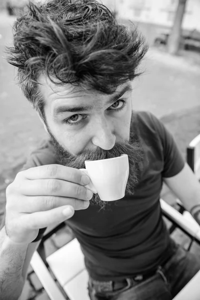 Hipster on surprised face drinking coffee outdoor. Man with beard and mustache holds cup of coffee while relaxing at cafe terrace. Coffee break concept. Guy having rest with espresso coffee
