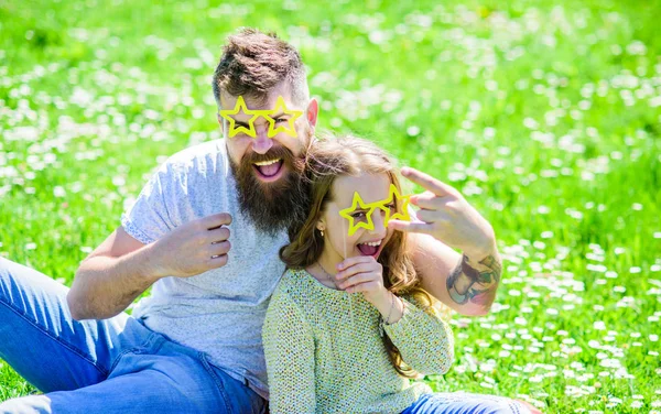 Rock star concept. Child and father posing with star shaped eyeglases photo booth attribute at meadow. Dad and daughter sits on grass at grassplot, green background. Family spend leisure outdoors