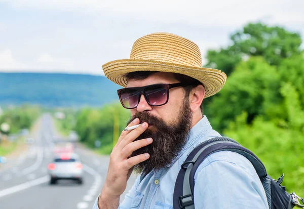 Cigarette before long journey. Traveler stylish hipster take brake.Man with beard and mustache in straw hat smoking cigarette, road background defocused. Smoking cigarette before long journey