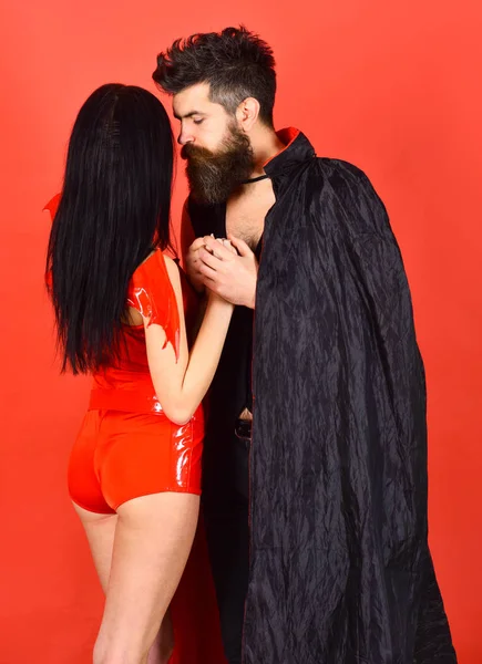 Vampire in cloak and sexy devil girl holds hands. Couple in love, perfect match. Couple on pensive faces play role game. Man and woman dressed like vampire, demon, red background. Halloween concept