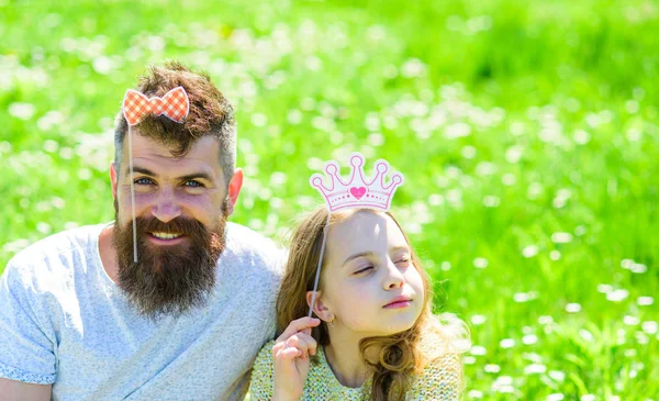 Family spend leisure outdoors, play girlish games. Child and father posing with crown and bow photo booth attributes. Dad and daughter sits on grass at grassplot, green background. Fatherhood concept — Stock Photo, Image