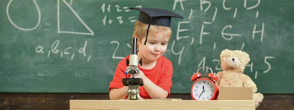 聪明的孩子的概念。孩子在愉快的面孔举行显微镜。首先对学习、学习、教育感兴趣。孩子在学术盖帽工作与显微镜在教室, 黑板在背景上 — 图库照片