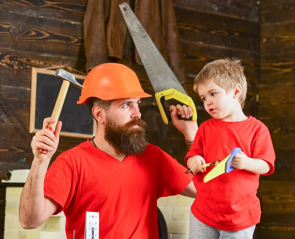 Babalık kavramı. Baba, üst koruyucu kask öğretim oğlu okul atölyesinde farklı araçları kullanmaya sakallı. Evlat, çocuk ciddi oyuncak testere, baba ile handcrafting sırasında eğlenmek tutar