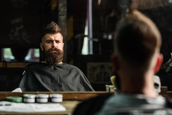 Hombre con barba cubierta con capa negro se sienta en la silla de peluquería delante del espejo. Hipster con barba comprobando el resultado del corte de pelo. Concepto de barbería. Hombre con barba cliente de barbería hipster — Foto de Stock