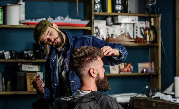 Hipster client with fresh haircut or hairstyle. Barber styling hair of bearded client with wax by hands. Barbershop concept. Man with beard and mustache in hairdressers chair, shelves on background — Stock Photo, Image