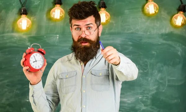 Der bärtige Hipster hält eine Uhr in der Hand, im Hintergrund eine Kreidetafel. Mann mit Bart und Schnurrbart im überraschten Gesicht. Lehrer mit Brille hält Wecker und zeigt mit Stift nach vorne. Disziplinkonzept — Stockfoto