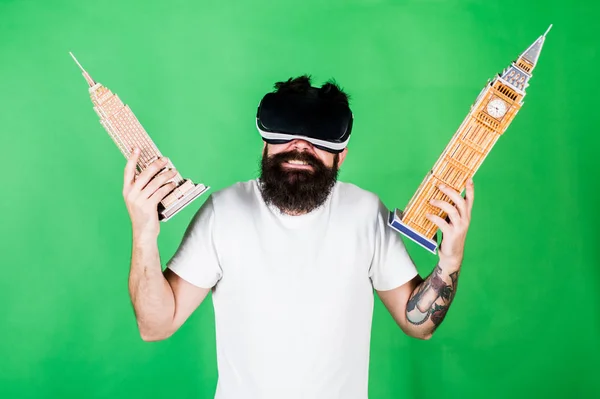 Man on confused face study architecture or design in virtual reality. 3D design concept. Man with beard in VR glasses, green background. Guy in VR glasses holds Big Ben and Empire State Building