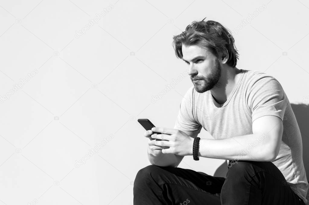 Man sit with smartphone on grey wall, new technology. Macho with beard, blond hair in tshirt on sunny day, fashion. New technology, mobile device. Mens fashion with accessory and style, copy space