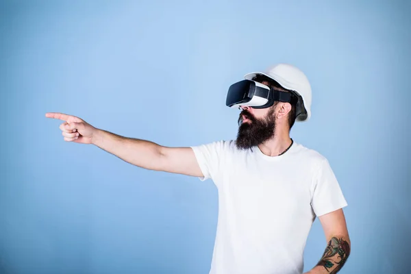 Hipster in helmet works as engineer in virtual reality. 3D design concept. Man with beard in VR glasses pointing with finger, light blue background. Architect or engineer with virtual reality glasses