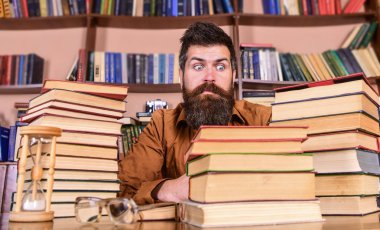 Öğretmen ya da öğrenci sakallı kum saati ve gözlük, defocused ile masaya otur. Kitap kurdu kavramı. Adam şok yüzündeki oturup Kitaplığı'nda, eğitim sırasında kitaplar, kitap rafı adam üzerinde yığınları arasında
