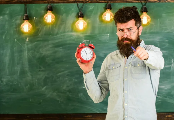 O hipster barbudo segura relógio, quadro no fundo, espaço de cópia. Homem com barba e bigode no rosto estrito. Professor em óculos detém despertador e apontando para a frente. Conceito de disciplina — Fotografia de Stock