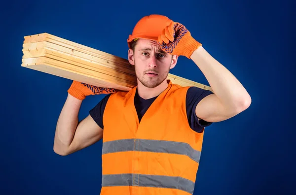 Плотник, столяр, рабочий, строитель несет деревянную балку на плече. Концепция деревообработки. Человек в защитных перчатках держит шлем, исправляет каску на голове, синий фон — стоковое фото