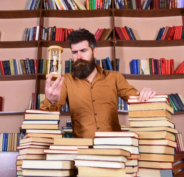 Könyvtáros koncepció. Tanár, tudós szakáll áll, könyvek, defocused asztalnál. Átgondolt arcán ember áll között könyvek, míg tart homokóra, halom könyvespolcok a háttérben — Stock Fotó