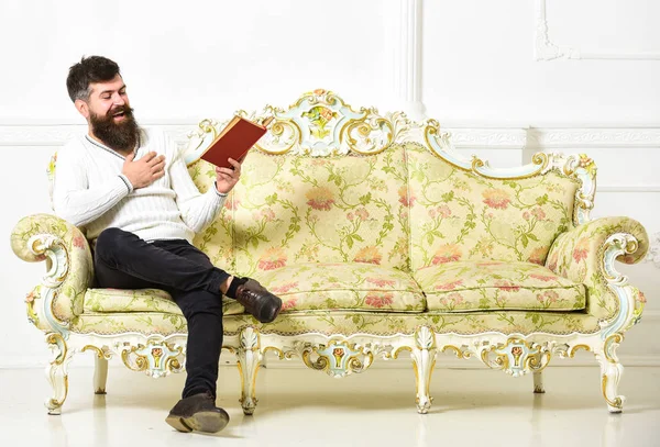 Un tipo leyendo un libro viejo con placer. Hombre con barba y bigote se sienta en el sofá de estilo barroco, sostiene libro, fondo blanco de la pared. Concepto de literatura humorística. Macho en la risa libro de lectura de la cara —  Fotos de Stock