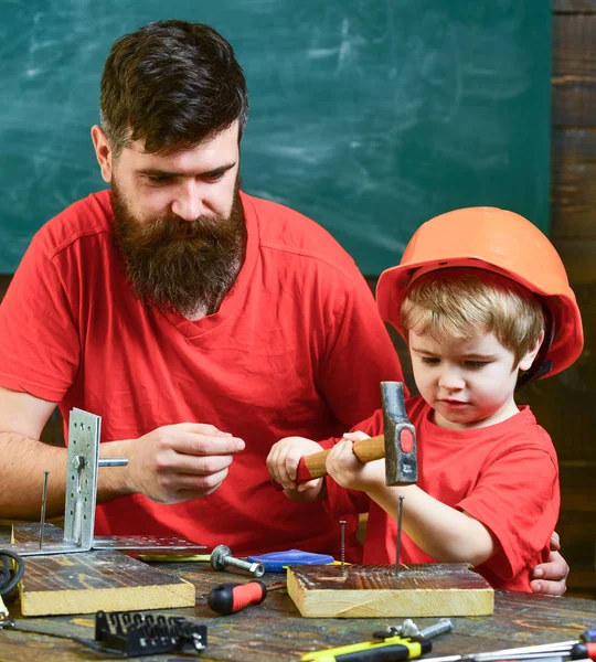 Babası öğretim araçları, çekiç, kullanmak için küçük oğlu sakal kara tahta arka plan ile. Çocuk, çocuk ile baba çekiç kullanmayı öğrenme koruyucu kask içinde meşgul. Tamirci kavramı