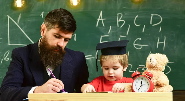 正式な摩耗と鏝板の教室で、背景に黒板に生徒の教師。少年は、するのに役立ちます、宿題のチェックの父の息子。ホーム スクーリングのコンセプトです。先生と勉強熱心な子供 — ストック写真