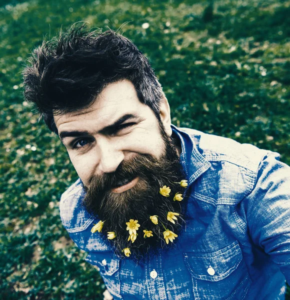 Hipster na cara piscando senta-se na grama, desfocado. Homem com barba gosta de primavera, fundo prado verde. Conceito da Primavera. Cara com flores menores de celandine na barba tirando foto selfie — Fotografia de Stock