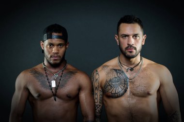 Ciddi yüz ve siyah arka plan üzerine izole uygun organları ile moda modelleri. Kartal dövme göğsünde geriye dönük şapkası ile Afrikalı adamın yanında ayakta beyaz adam ile geometrik dövmeler