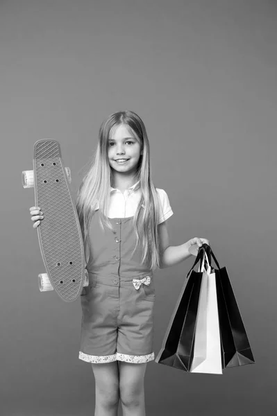 Chica feliz con tabla de skate y bolsas de compras sobre fondo violeta. Niño pequeño sonríe con bolsas de papel y monopatín. Después de ir de compras. venta y viernes negro. Deporte y juegos activos —  Fotos de Stock