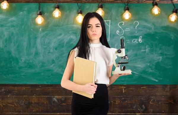 Professeur de biologie détient livre et microscope. Concept de biologie. Lady scientifique tient livre et microscope, tableau sur fond, espace de copie. Dame en tenue formelle sur le visage calme en classe — Photo