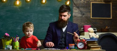 Sanat ders kavramı. Sakallı öğretmeni, sınıfta, kara tahta arka plan çizmek için küçük oğlu baba öğretir. Çocuk ve meşgul yüz boyama, resim öğretmeni. Yetenekli sanatçı oğlumla vakit geçirmek