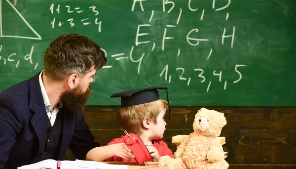 個人レッスンのコンセプト。先生が説明する間、男の子、大学院生の帽子の子供は黒板の落書きを見ています。ひげを生やした教師、父は教室で小さな息子を教えています。 — ストック写真