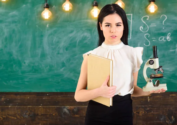 Concept de biologie. Professeur de biologie détient livre et microscope. Dame en tenue de cérémonie sur le visage calme en classe. Lady scientist tient livre et microscope, tableau sur fond, espace de copie — Photo