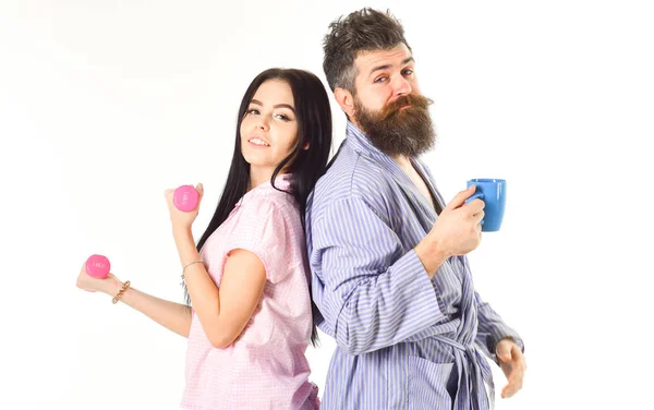 Alternative lifestyle concept. Couple, family on sleepy faces, full of energy. Couple in love in pajama, bathrobe stand isolated on white background. Girl with dumbbell, man with coffee cup