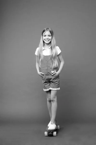 Menina pequena sorriso com skate bordo no fundo violeta. Patinador infantil com longboard. Miúdo de skate de fato-macaco rosa. Atividades desportivas e energia. Patinar é a vida deles. Infância e jogos ativos — Fotografia de Stock