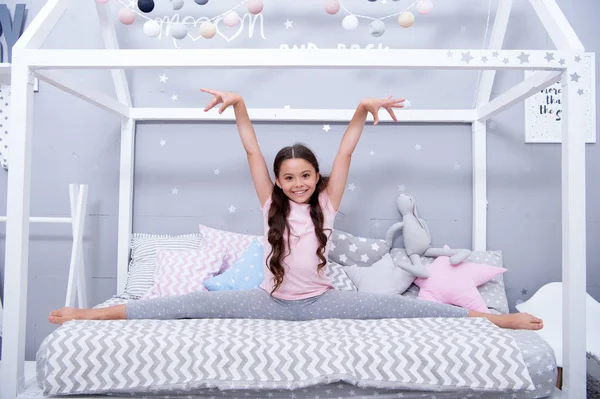 Cute gymnast practice split before go to bed. Girl child sit split bed in her bedroom. Kid prepare to go to bed. Time for evening stretching. Girl kid long hair cute pajamas relaxing and stretching