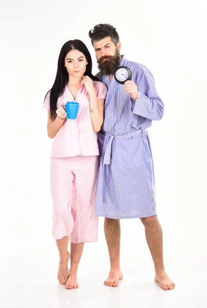 Perfekt morgon koncept. Par, familj vaknade i tid. Par i kärlek, unga familj i pyjamas, morgonrock stå isolerade på vit bakgrund. Flicka med kaffekoppen, man håller klockan i hand — Stockfoto