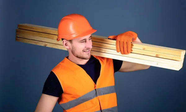 Man in de helm, helm en beschermende handschoenen houdt houten balken, grijze achtergrond. Timmerman, schrijnwerker, arbeider, bouwer op het glimlachen gezicht draagt houten balken op de schouder. Hardy arbeider concept — Stockfoto