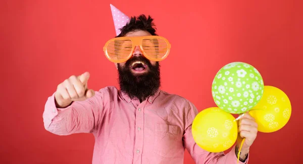 Hipster in gigantische zonnebril viert verjaardag. Man met baard en snor op blij gezicht houdt stelletje lucht ballonnen, rode achtergrond. Guy in feest hoed wijzen naar voren. Verjaardag partij concept — Stockfoto