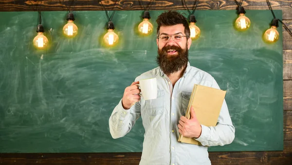 Professor em óculos detém livro e caneca de café ou chá. Conceito de pausa para café. Homem com barba na cara feliz na sala de aula. Cientista detém livro e caneca de café, quadro no fundo, cópia spac — Fotografia de Stock