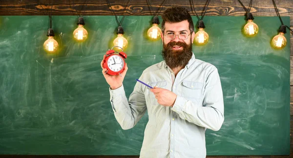 O hipster barbudo segura relógio, quadro no fundo, espaço de cópia. Homem com barba e bigode no rosto sorridente fica na sala de aula. Conceito de disciplina. Professor em óculos detém despertador e caneta — Fotografia de Stock