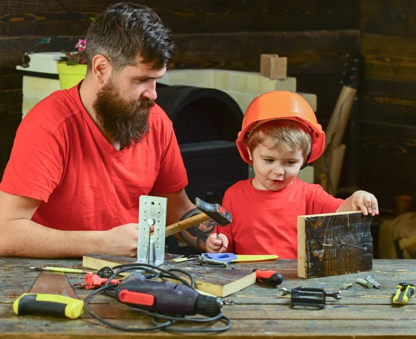 Fatherhood concept. Father with beard teaching little son to use tools, hammering, chalkboard on background. Boy, child busy in protective helmet learning to use hammer with dad.