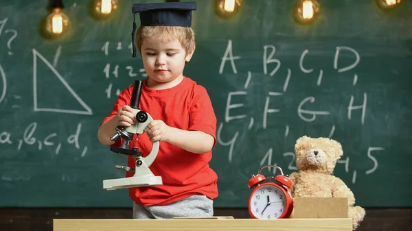 Primeiro ex-interessado em estudar, aprender, educação. Criança no rosto ocupado perto do microscópio. Conceito de criança inteligente. Kid boy em trabalho cap acadêmico com microscópio em sala de aula, quadro-negro em segundo plano — Fotografia de Stock