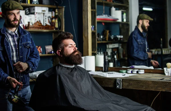 Hipster bearded client got hairstyle. Barber with hairdryer blows off hair out of cape. Barber with hairdryer works on hairstyle for bearded man, barbershop background. Barbershop concept — Stock Photo, Image