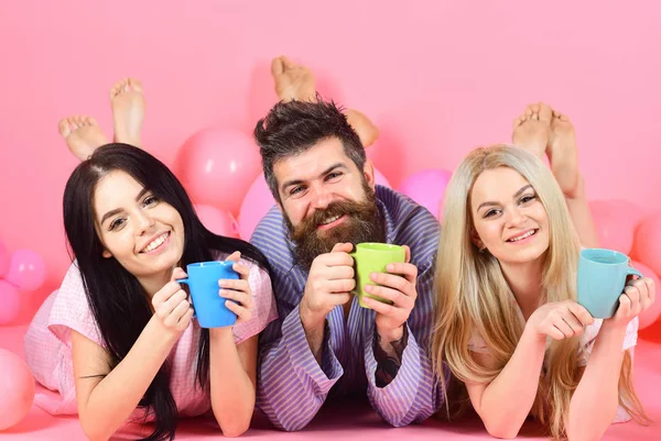 Lovers drinking coffee in bed. Man and women in domestic clothes, pajamas. Man and women, friends on happy faces lay, pink background. Threesome relax in morning with coffee. Lovers in bed concept