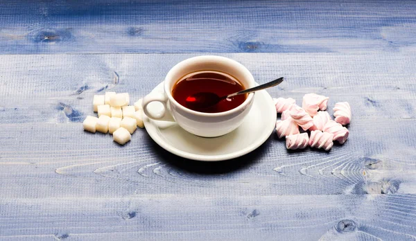お茶の時間の概念。飲み物とお菓子のデザートとして用意します。マグカップは、入れたての紅茶、スプーンとマシュマロと青い木製の背景に洗練された砂糖の山でいっぱい。紅茶とお菓子とマグカップ — ストック写真
