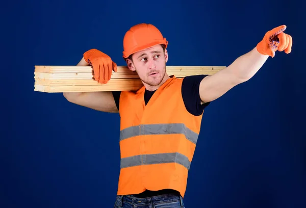 Holzwerkstoffkonzept. Tischler, Holzarbeiter, kräftiger Bauarbeiter auf dem Gesicht trägt Holzbalken auf der Schulter. Mann mit Helm, Schirmmütze und Schutzhandschuhen weist Richtung, blauer Hintergrund — Stockfoto
