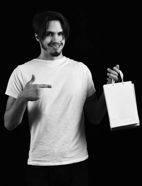 Beyaz tişörtlü yakışıklı, şık saçlı, yüzünde renkli bir paket ya da siyah stüdyo arka planında çantalar olan bir adam.