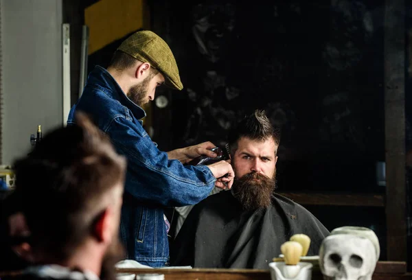 Hipster client getting haircut. Haircut concept. Barber styling hair of brutal bearded client with clipper. Barber with hair clipper works on hairstyle for man with beard, barbershop background — Stock Photo, Image
