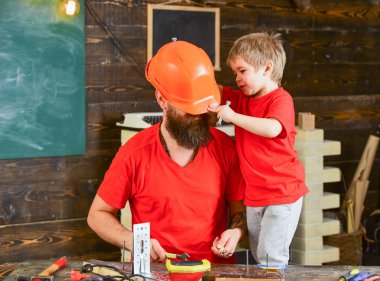 Baba, ana okul atölyesinde farklı araçları kullanmak için kask öğretim oğlu sakallı. Evlat, çocuk neşeli cıvata veya vida, baba ile handcrafting sırasında eğlenmek tutar. Tamirci kavramı