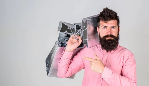 Concepto de recomendación. Un tipo de compras en temporada de ventas, apuntando a las bolsas. El hombre con barba y bigote lleva un montón de bolsas de compras, fondo gris. Hipster en la cara seria recomienda comprar — Foto de Stock