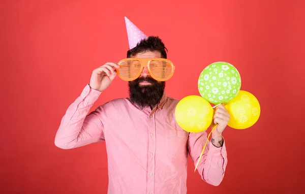 Хіпстер у гігантських сонцезахисних окулярах, що святкують день народження. Чоловік з бородою і вусами на щасливому обличчі тримає купу повітряних кульок, червоний фон. Концепція святкування. Хлопець у вечірньому капелюсі святкує свято — стокове фото