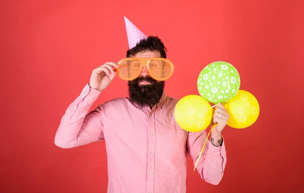 Hipster em óculos de sol gigantes comemorando aniversário. Homem com barba e bigode na cara feliz segura um monte de balões de ar, fundo vermelho. Conceito de celebração. Cara em chapéu de festa celebra férias — Fotografia de Stock