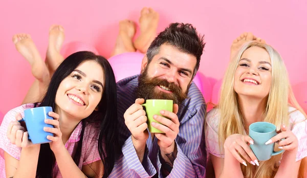 Liebhaber trinken Kaffee im Bett. Mann und Frau, Freunde auf lächelnden Gesichtern, rosa Hintergrund. Männer und Frauen in Hauskleidung, Schlafanzug. Drei entspannen am Morgen mit Kaffee. Liebespaar im Bett — Stockfoto