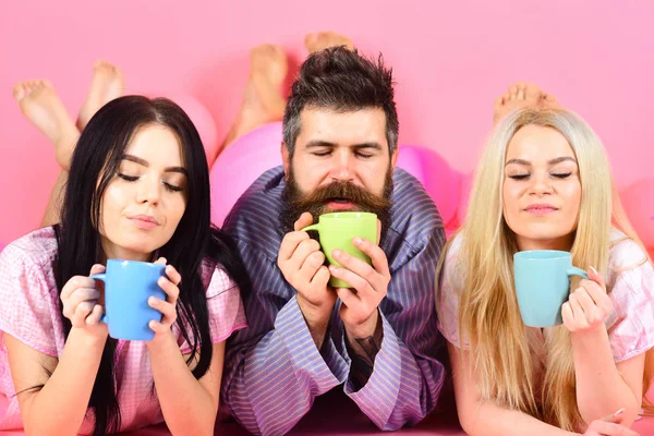 Lovers drinking coffee in bed. Threesome relax in morning with coffee. Stormy night concept. Man and women in domestic clothes, pajamas. Man and women, friends on sleepy faces lay, pink background