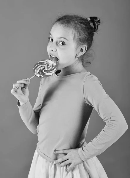 Child with curious face poses with candy on green background. Girl eats big colorful sweet caramel. Treatment and sweets concept.
